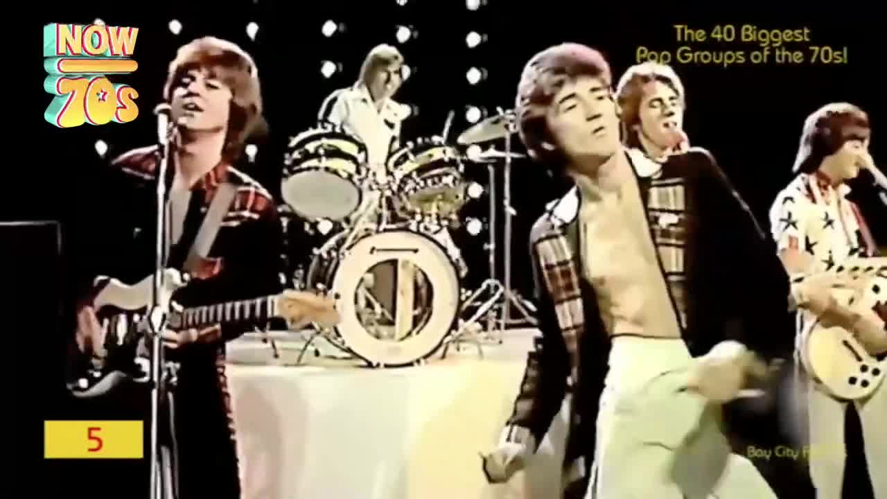 A band with matching plaid shirts plays on a stage, the shirtless singer moves to the music. The drummer keeps the beat, the guitarists strum, and the crowd is loving it.
