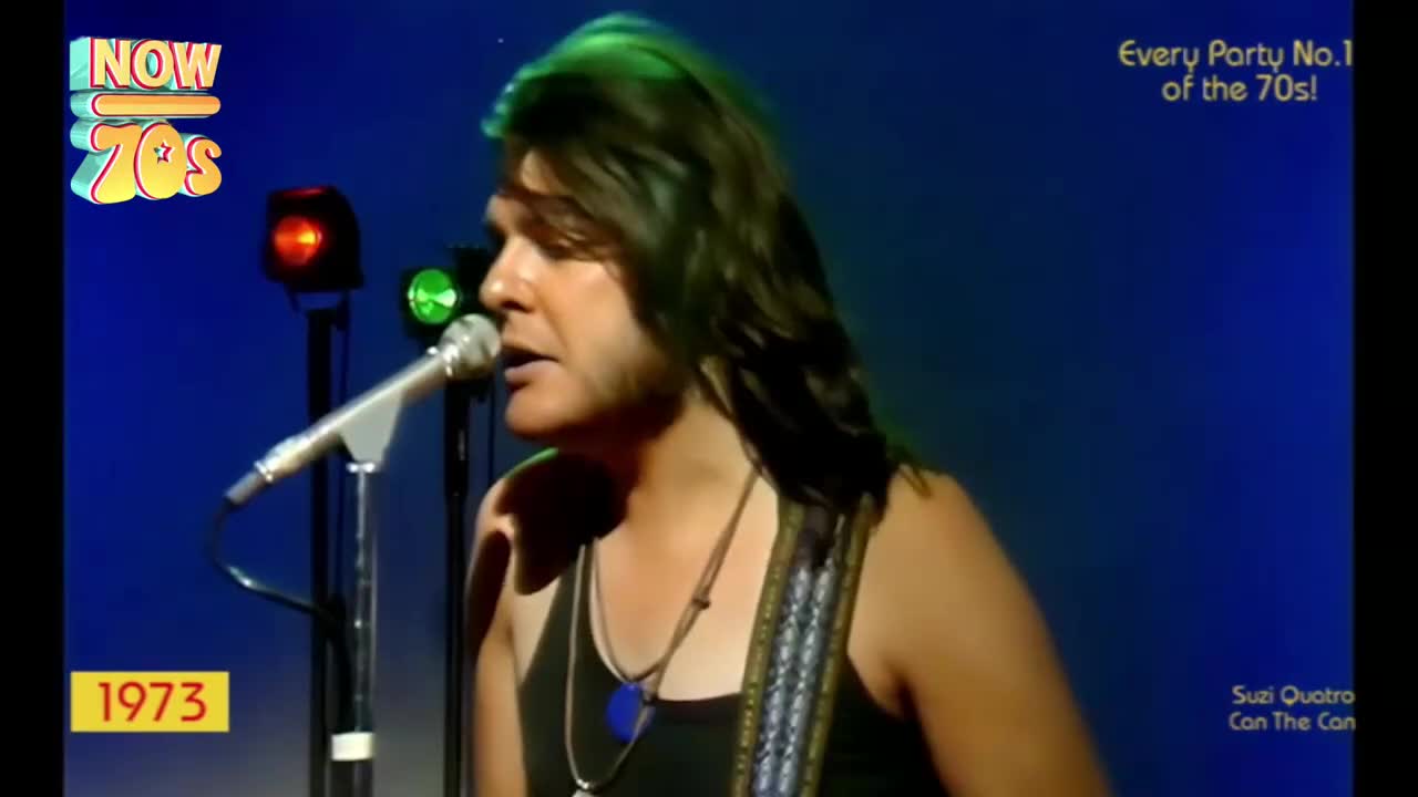 A man with long hair, wearing a tank top and a guitar strap, sings into a microphone. The stage lights cast a green glow on his hair as he performs.
