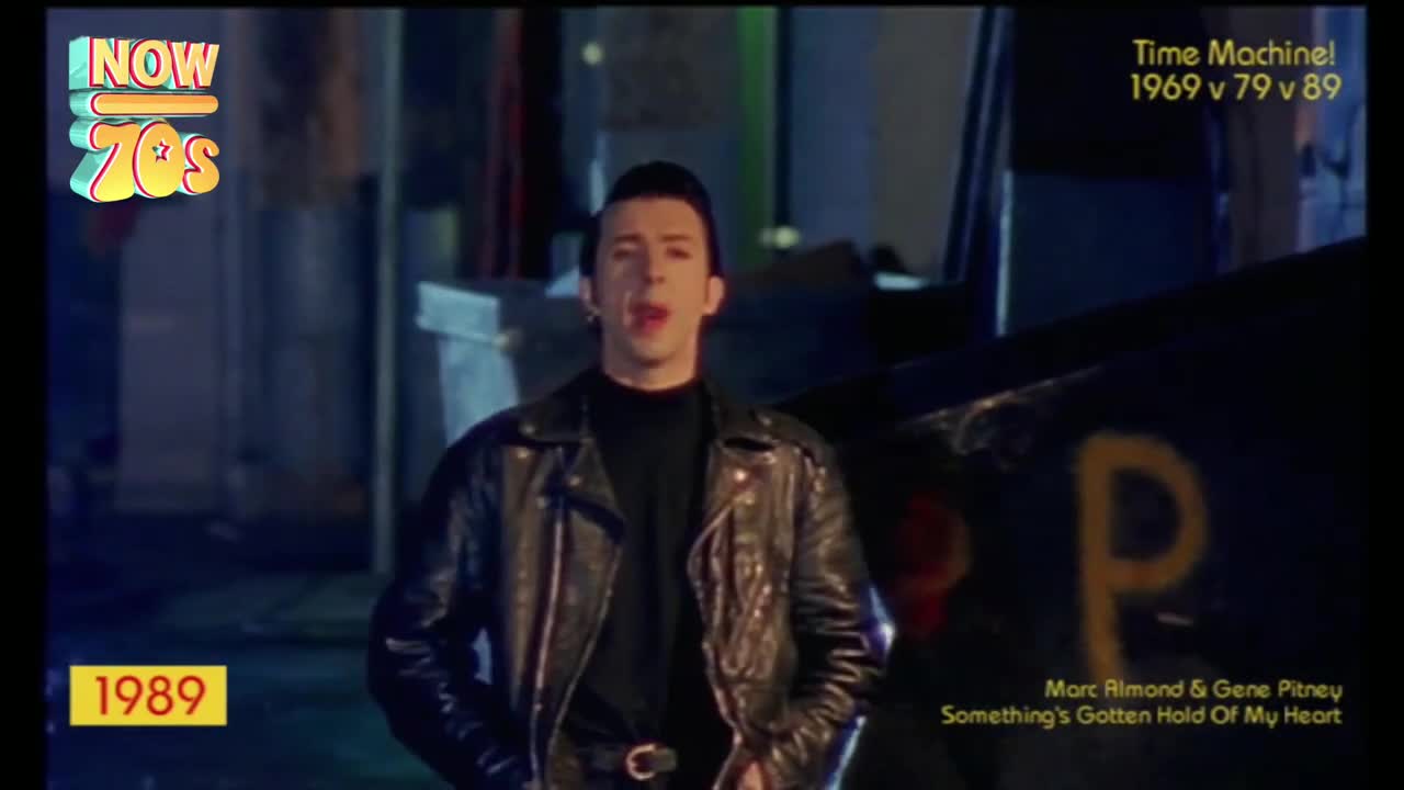 A man in a leather jacket is singing, his mouth open as he faces the camera. The backdrop is a dark, industrial-looking scene, with a large "P" painted on something behind him.
