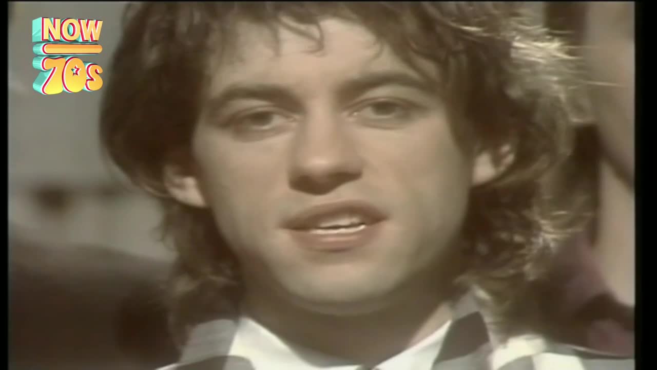 A man with wavy hair and a checkered shirt is speaking directly to the camera. The "Now 70s" logo sits in the corner, a reminder of the era.
