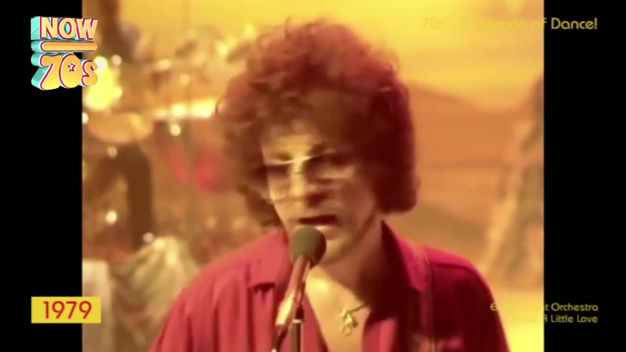 The singer, in a red shirt, is holding a microphone, his curly hair framing his face. Behind him, the band is barely visible in the warm stage lighting.
