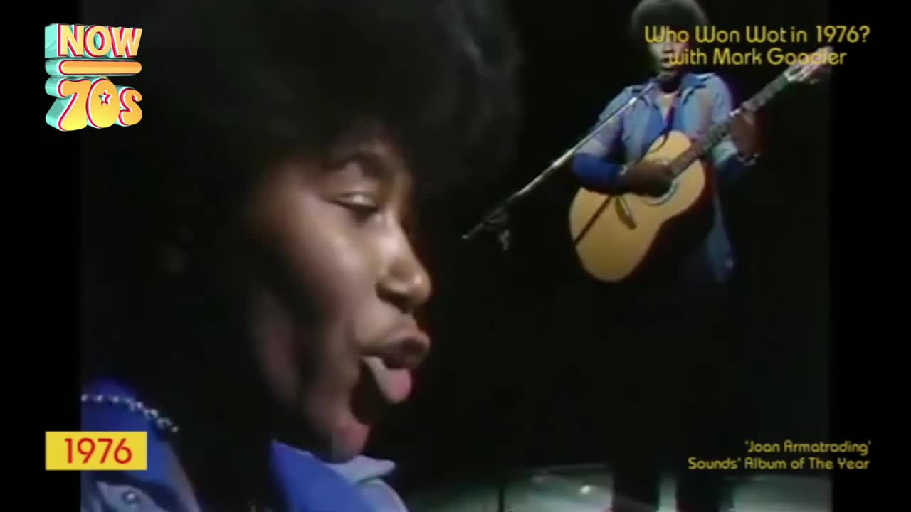 A woman with dark hair is singing, her mouth wide open. Behind her, a man strums an acoustic guitar.
