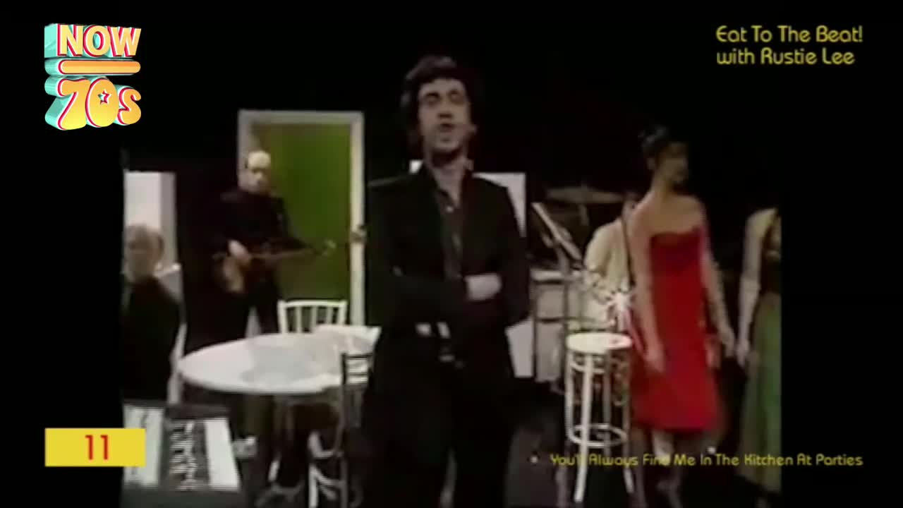 A man in a dark suit stands center stage, singing with his arms crossed. A woman in a bright red dress moves to his right, and a band plays behind them.
