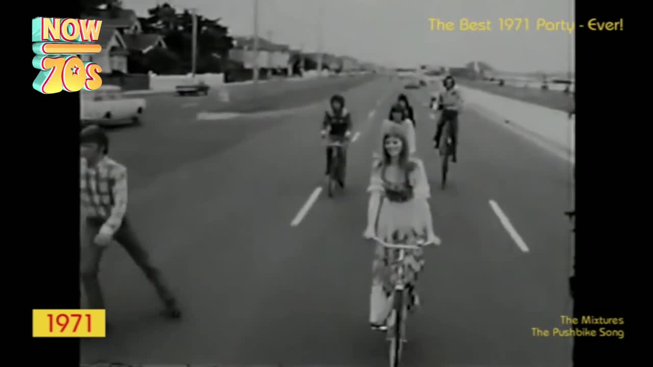 A group of people are cycling down a road, the woman in front smiling at the camera. A man walks alongside them, dressed in a checked shirt and jeans.
