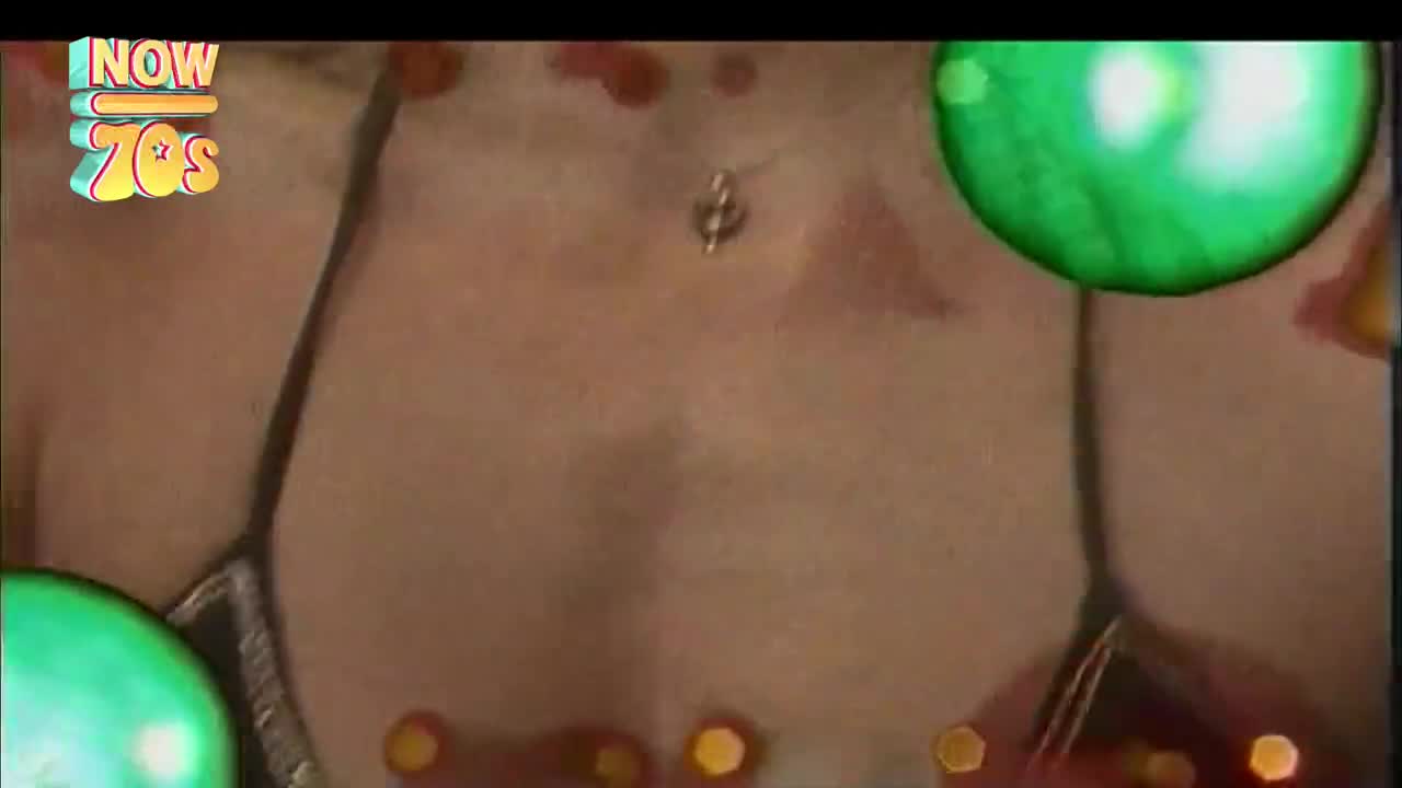 A torso is framed by a dark, glittering garment. Green orbs and small, golden shapes float around the body.
