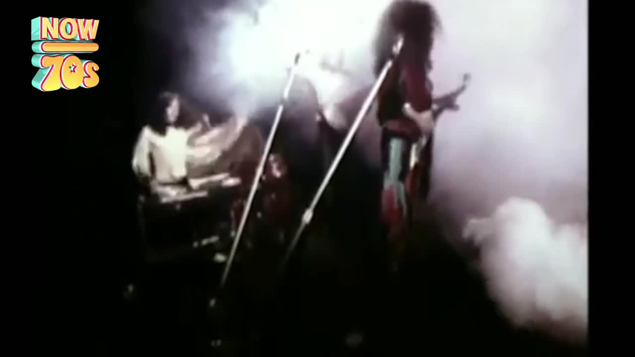 A band is bathed in a haze of stage smoke, the guitarist in a red and black outfit. The keyboardist is hunched over his instrument, while the guitarist's long hair whips around as he plays.
