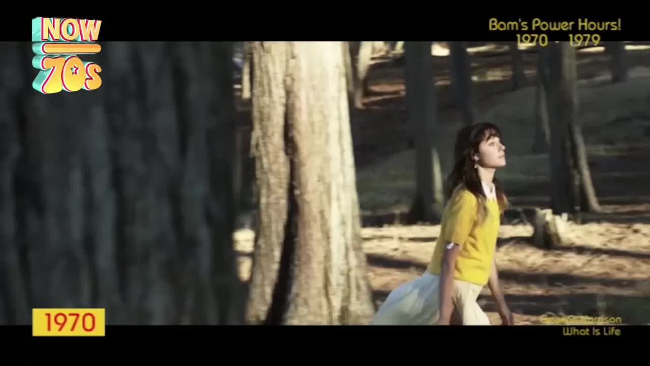 A young woman in a yellow top and white skirt walks through a sun-dappled forest, looking upwards. The trees stand tall and the light catches her hair as she moves.
