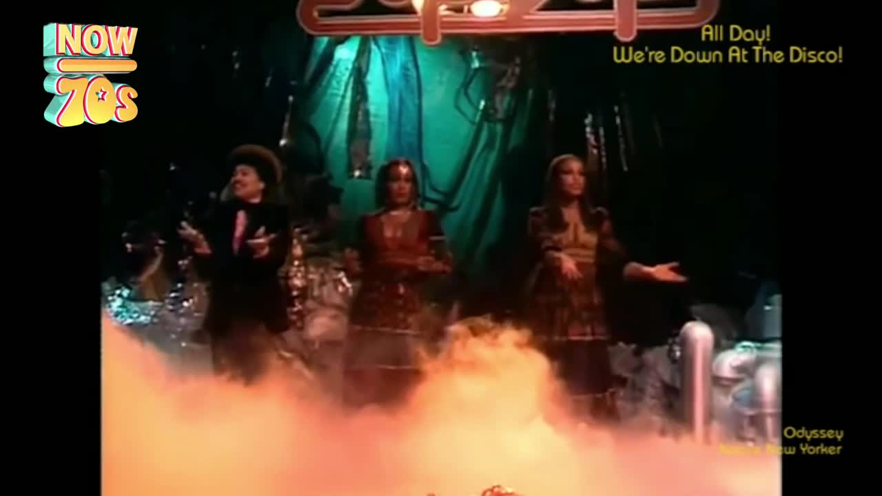Three performers, dressed in glittery outfits, stand on a stage shrouded in a thick, orange fog. They're gesturing with their hands, and a backdrop of shimmering fabric hangs behind them.
