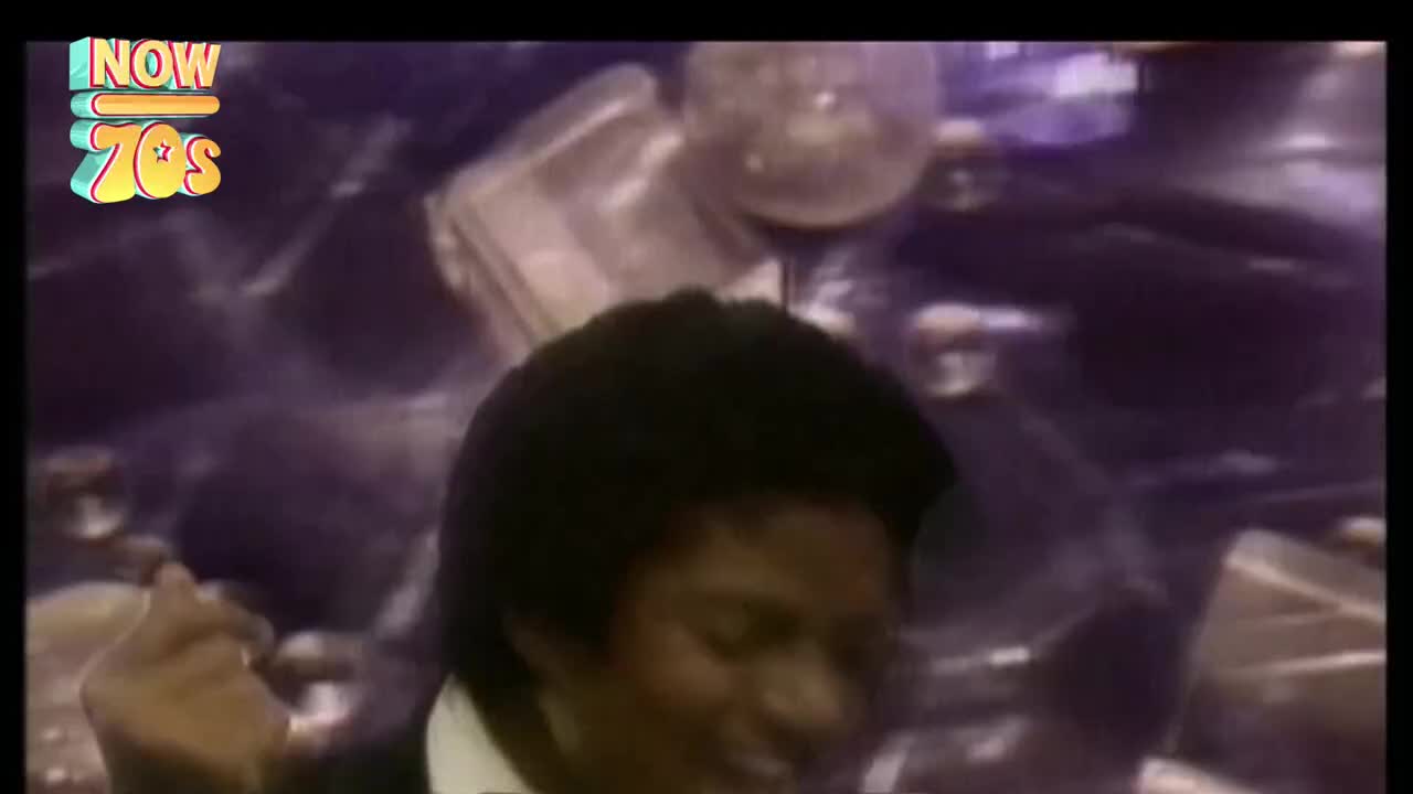 The singer, hair slicked back, is grooving, his arm up in the air. Behind him, a sea of purple and silver shapes shimmers under the stage lights.
