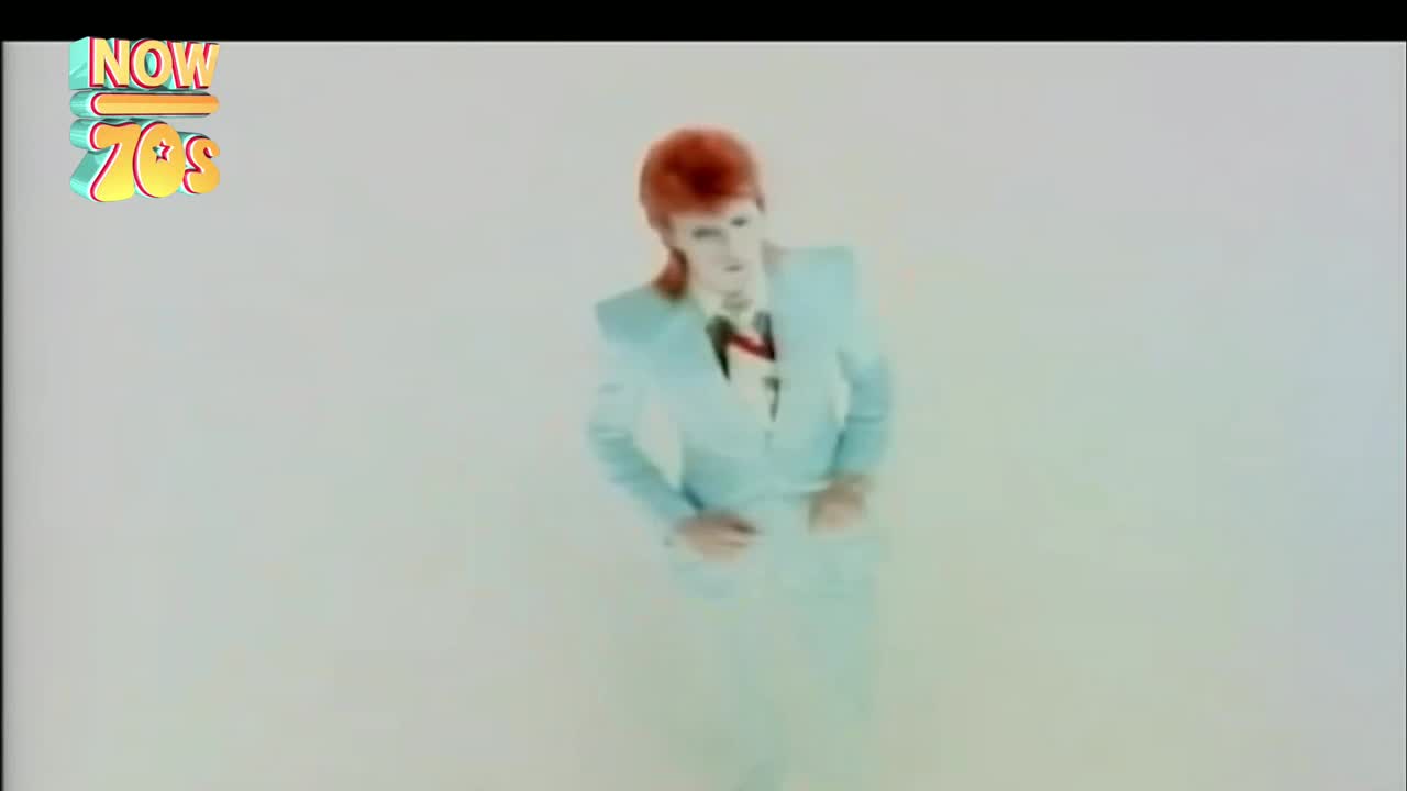 He's standing center stage, in a pale blue suit with a red-haired mullet. The backdrop is a hazy white, and he's looking straight ahead.
