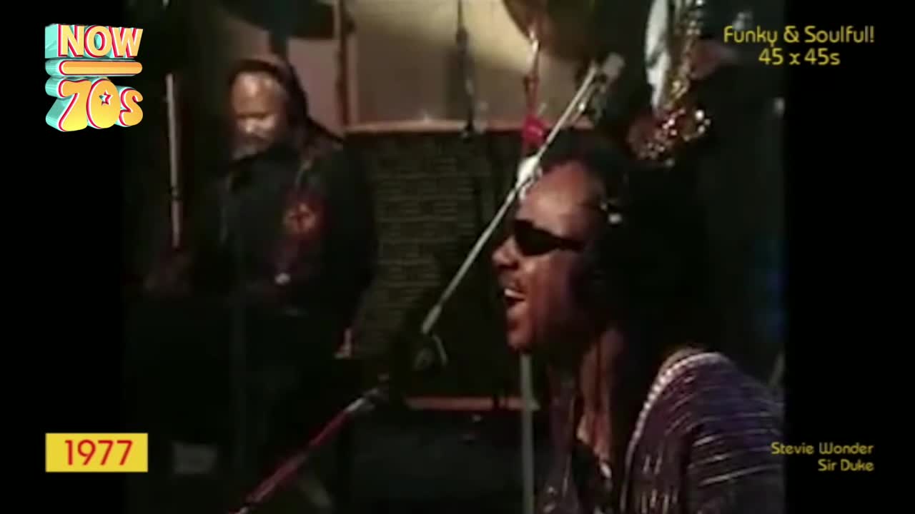 Stevie Wonder, in dark shades and a patterned shirt, sings into a microphone, his face lit up. Behind him, a band member stands, a saxophone glinting in the studio lights.
