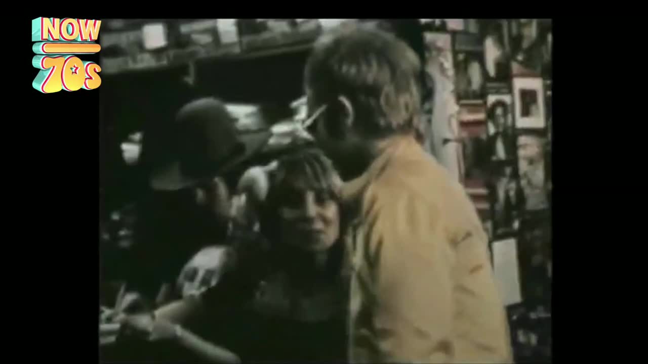 Someone in a yellow shirt is leaning in, hand on a woman's shoulder, as they chat. Behind them, a wall of posters and a man in a hat are visible in the dimly lit space.
Someone in a yellow shirt is leaning in, hand on a woman's shoulder, as they chat. Behind them, a wall of posters and a man in a hat are visible in the dimly lit space.