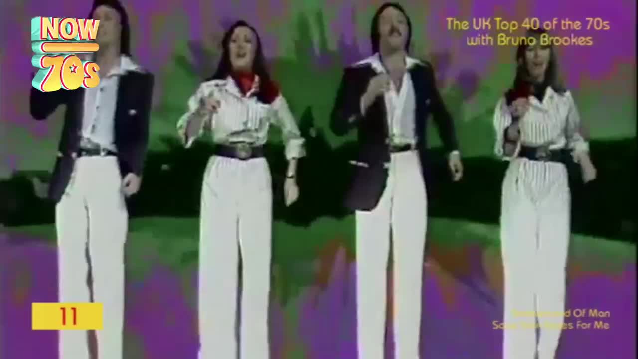 Four people, dressed in white trousers and open shirts, are singing and swaying on a brightly colored stage. The men wear black jackets, while the women have scarves around their necks, all performing on a show from the UK.
Four people, dressed in white trousers and open shirts, are singing and swaying on a brightly colored stage. The men wear black jackets, while the women have scarves around their necks, all performing on a show from the UK.