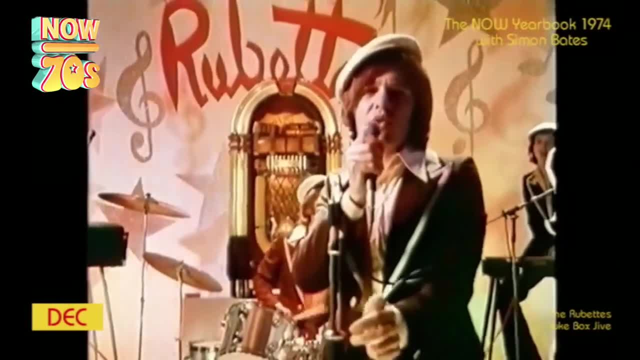 The lead singer, in a cap and suit, belts into the microphone, backed by a band playing in front of a jukebox. The stage is decorated with musical notes and the band's name, "Rubettes," in a retro font.
The lead singer, in a cap and suit, belts into the microphone, backed by a band playing in front of a jukebox. The stage is decorated with musical notes and the band's name, "Rubettes," in a retro font.
