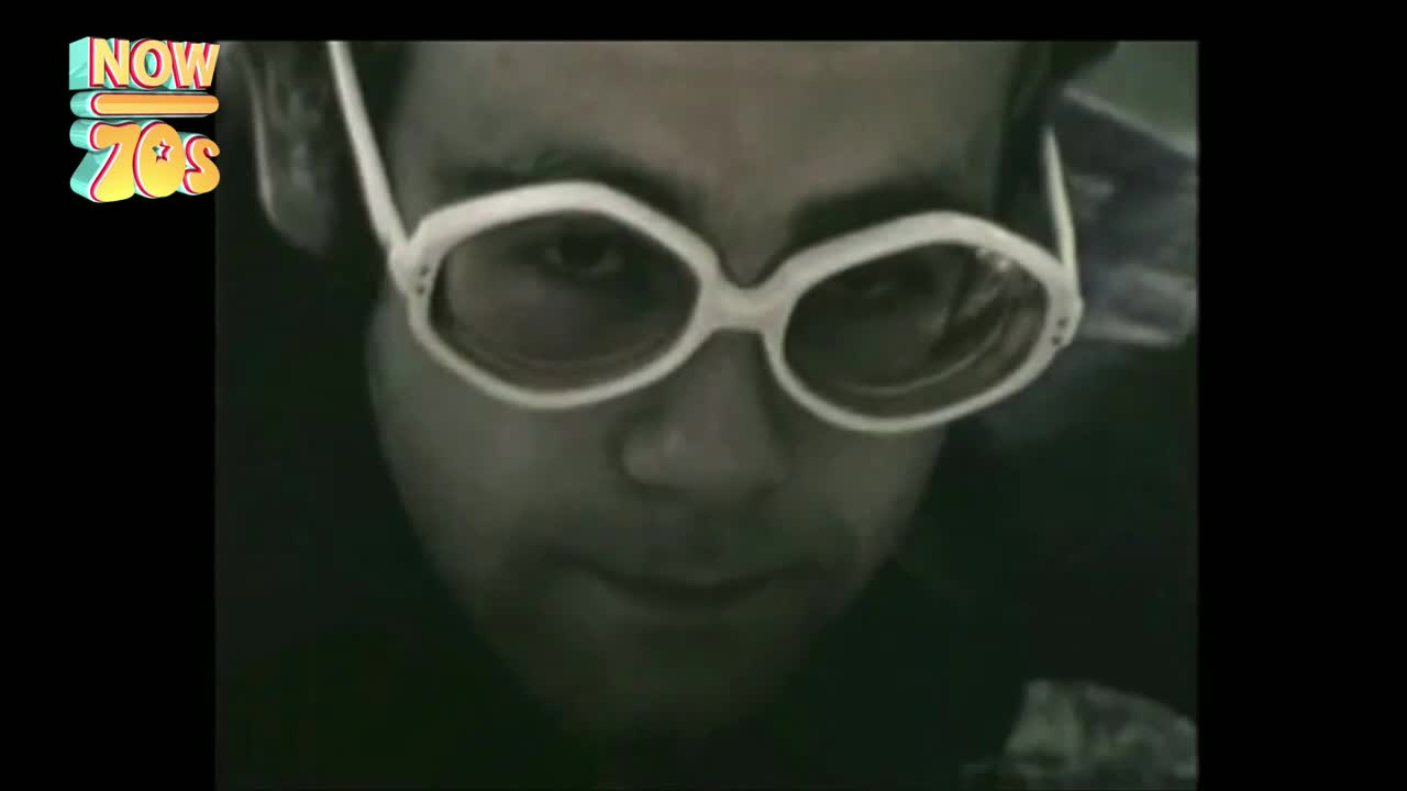 A man with oversized white-rimmed glasses stares directly ahead. The monochrome image and the retro logo in the corner suggest this is a clip from the UK, Now 70s.
A man with oversized white-rimmed glasses stares directly ahead. The monochrome image and the retro logo in the corner suggest this is a clip from the UK, Now 70s.