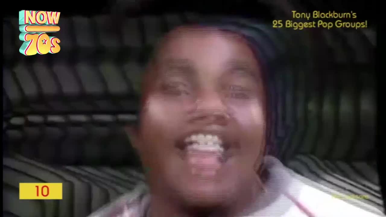 A smiling face beams from the screen, framed by a striped background. The "Now 70s" logo and a countdown number are visible in the corner.
A smiling face beams from the screen, framed by a striped background. The "Now 70s" logo and a countdown number are visible in the corner.