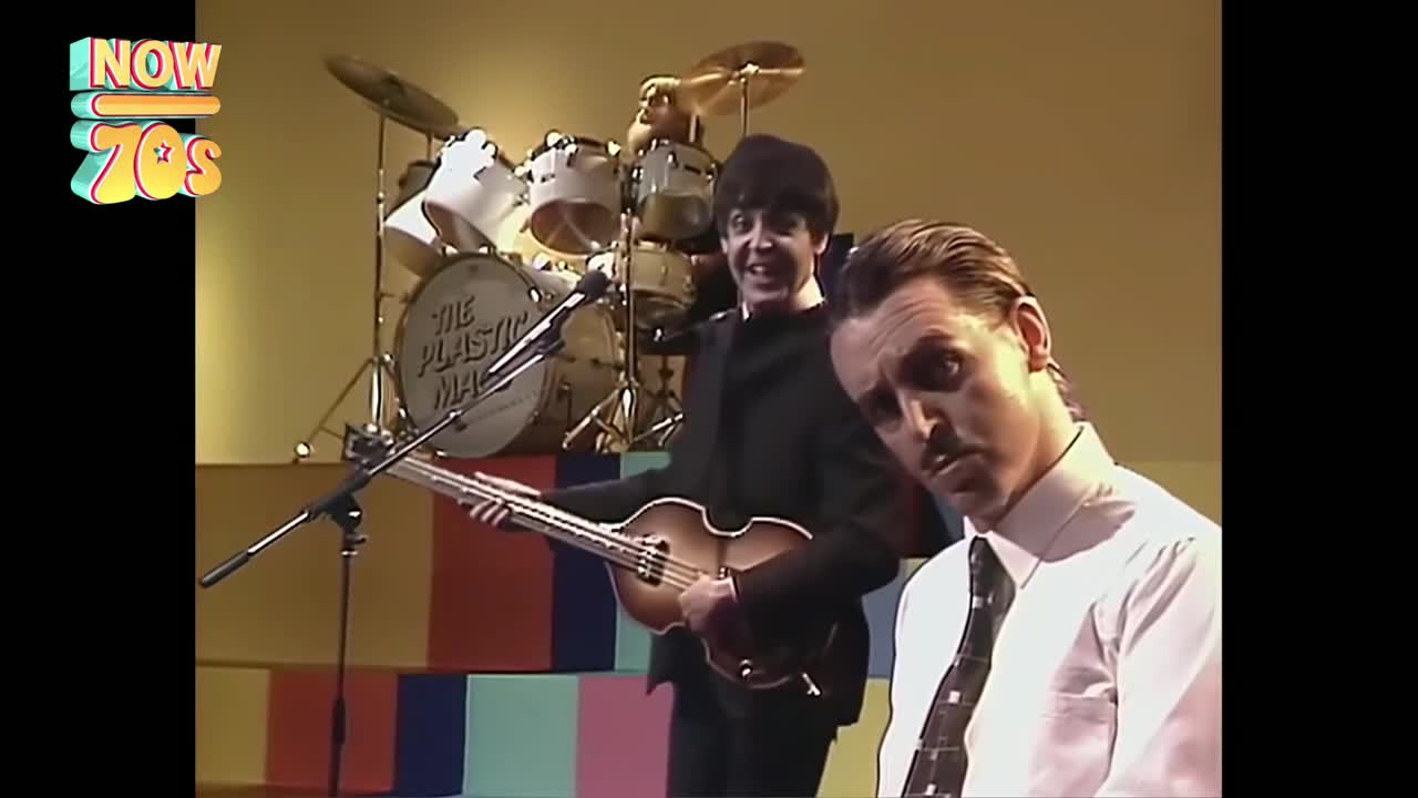 Paul McCartney, grinning, strums a Hofner bass guitar while a man in a tie looks directly at the camera. Behind them, a drum kit with "The Plastic Man" on the bass drum sits ready.
Paul McCartney, grinning, strums a Hofner bass guitar while a man in a tie looks directly at the camera. Behind them, a drum kit with "The Plastic Man" on the bass drum sits ready.