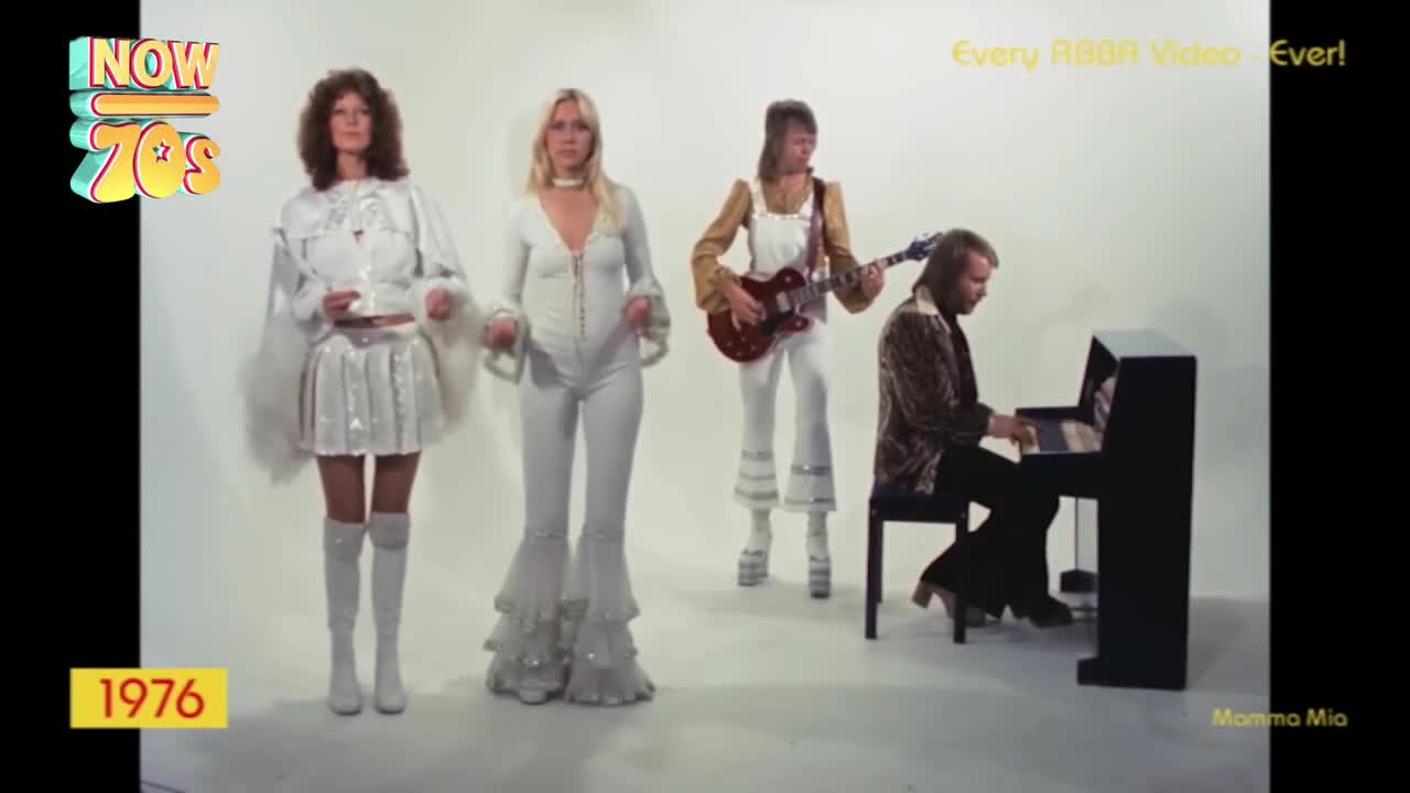 Four people are in a stark white space, the women in white outfits with flared sleeves and pants, while the men play guitar and piano. The band is performing, with the piano player seated and the others standing, all bathed in bright light.
