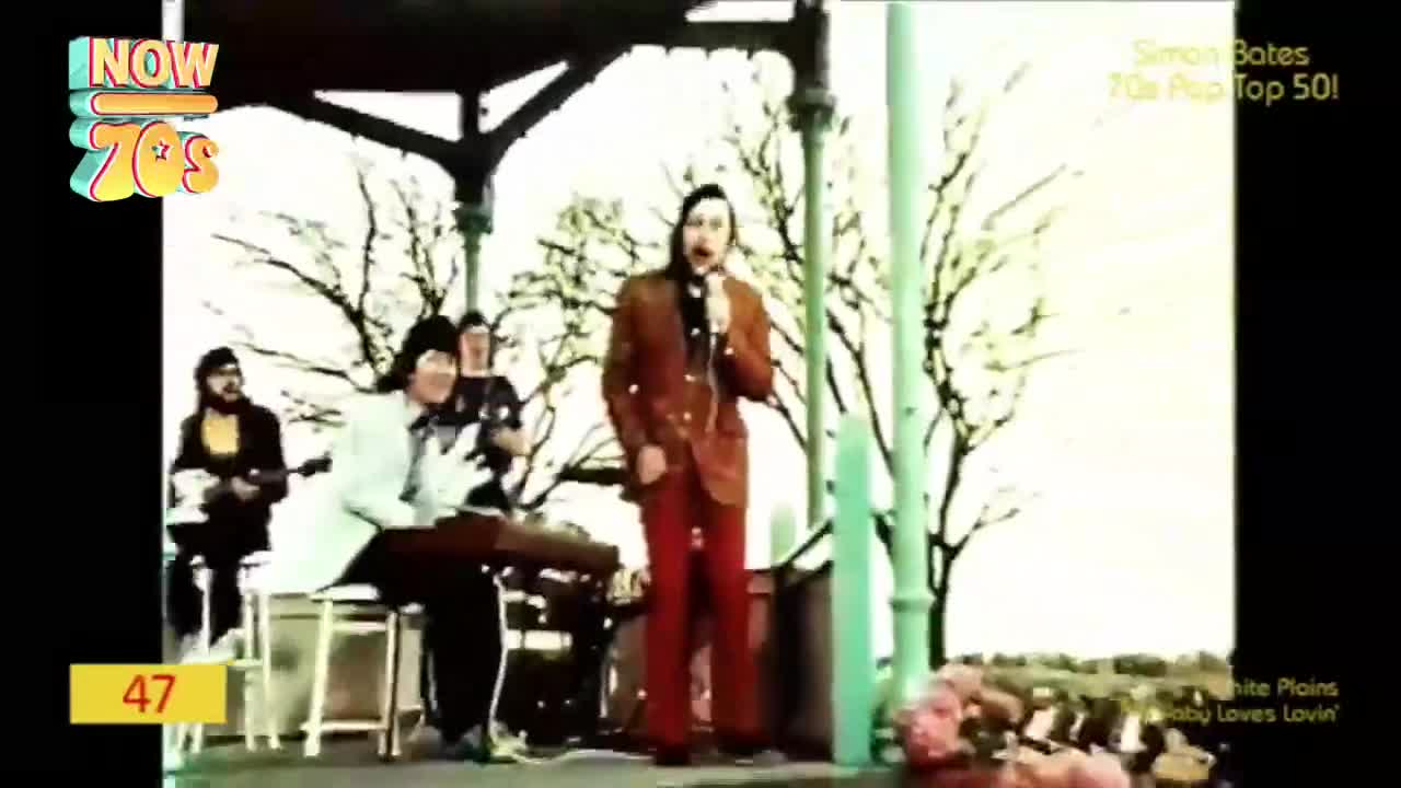The singer in the brown jacket and red trousers belts into the microphone, his band backing him on a stage. The keyboardist in the white suit is seated, and the drummer is visible behind him.
