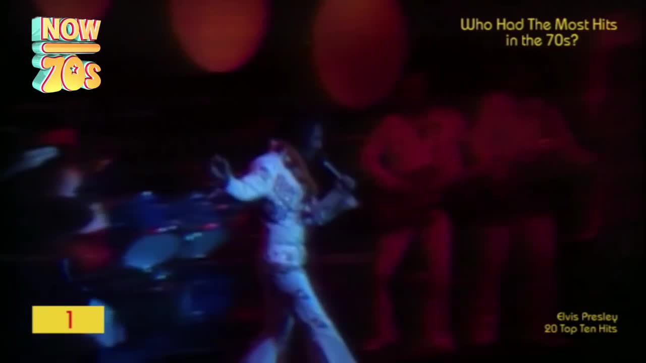 A man in a white jumpsuit, holding a microphone, is center stage. Behind him, two figures in silhouette move to the music.
