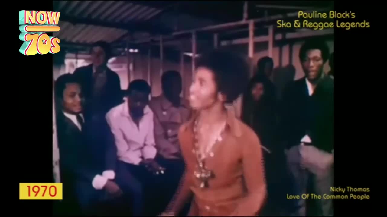 A woman with a large afro and a long necklace sings into a microphone, her eyes closed. Several men in suits and shirts stand and sit behind her, watching the performance.
