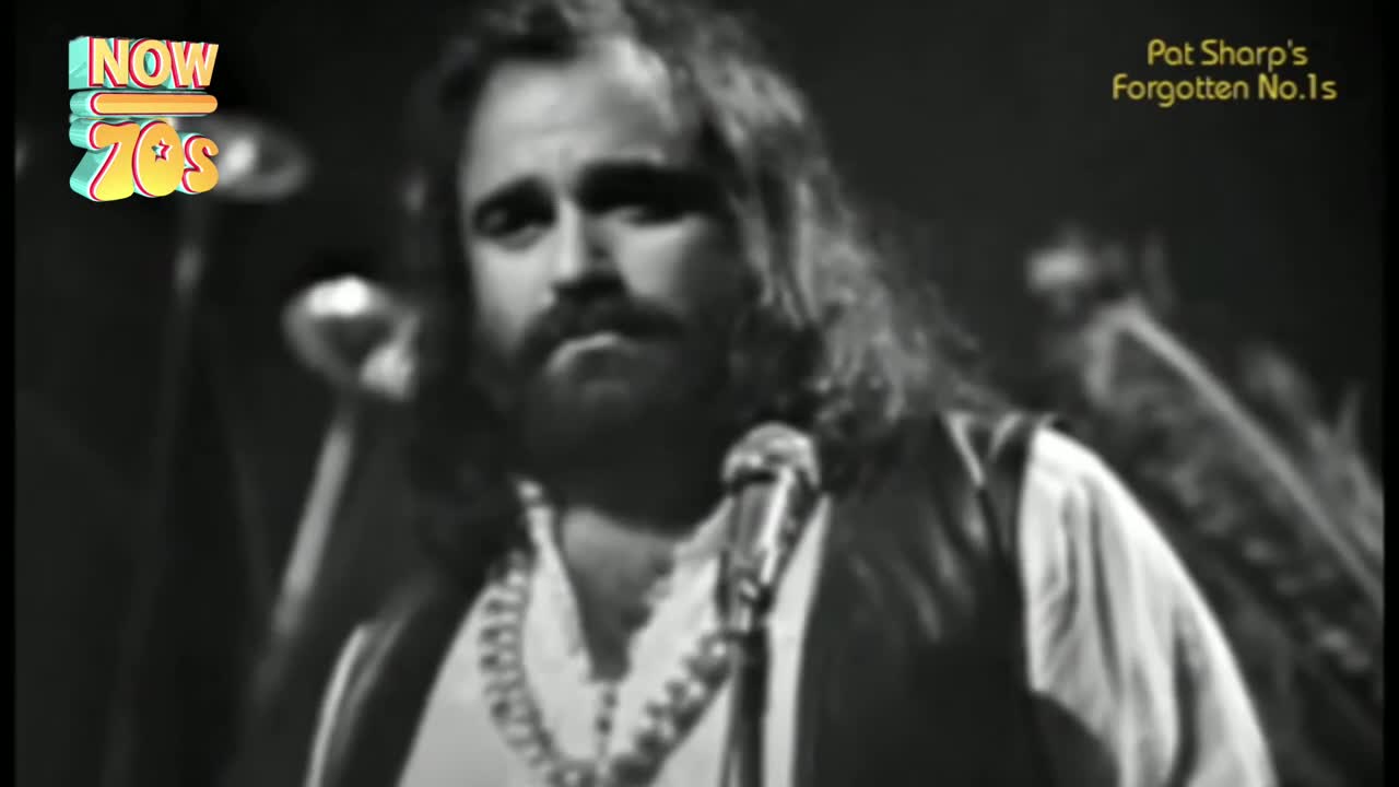 The singer, with long hair and a beard, stands at the microphone, looking directly at the camera. He's wearing a shirt and a vest, and a necklace hangs around his neck.
