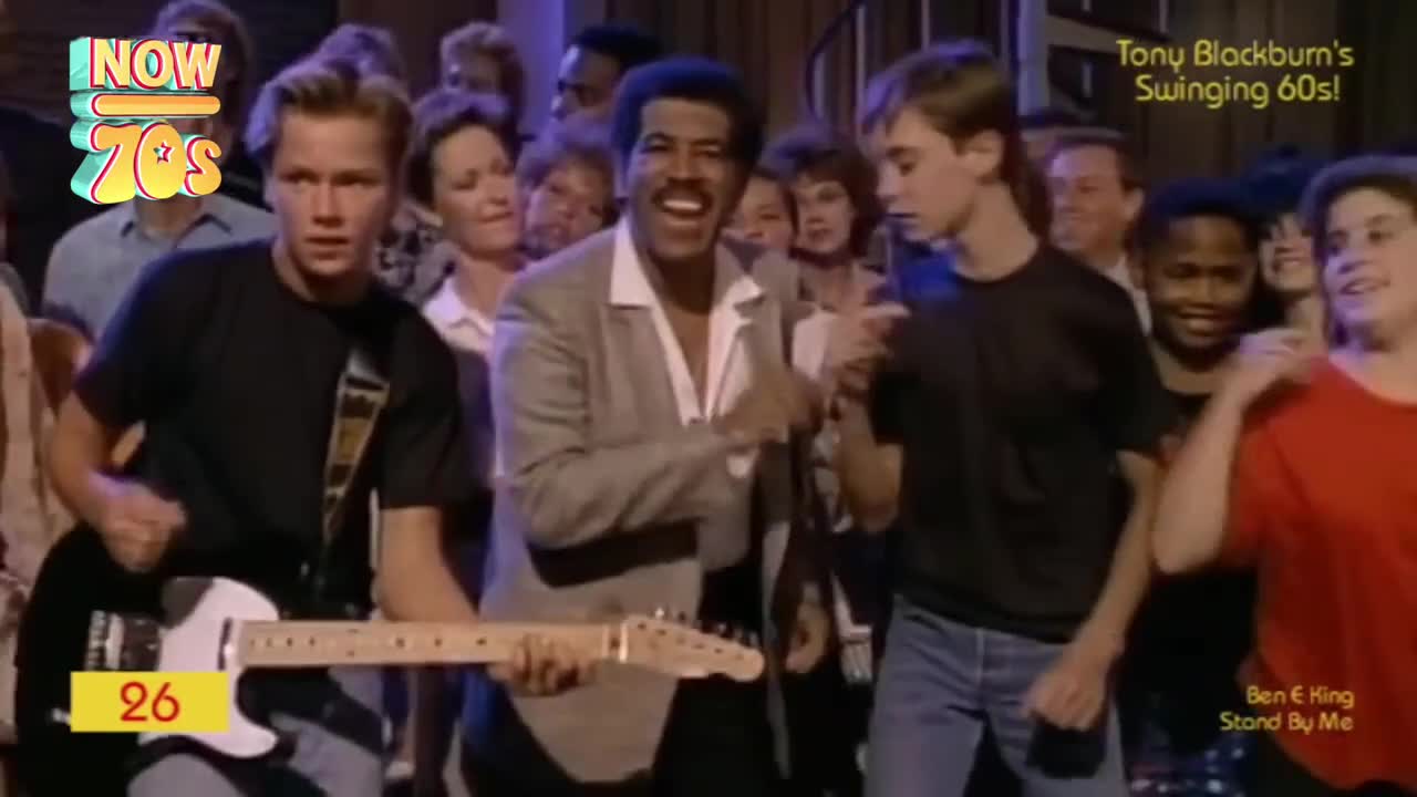 A man in a light suit grins, singing into the camera as the crowd sways to the music.  A guitarist strums a white Fender, while the audience dances along to a familiar tune.
