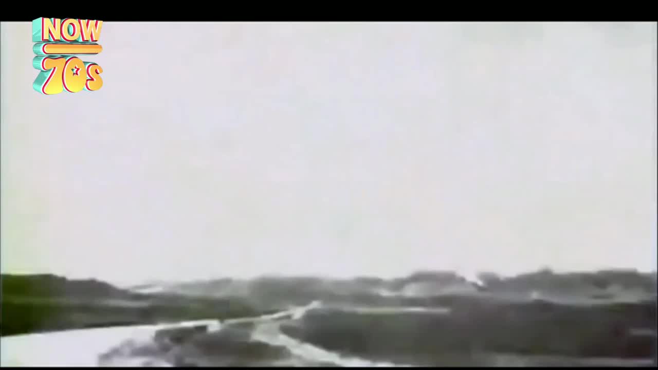 The train is speeding along the tracks, its carriages a dark shape against the bright sky. The landscape rushes past, a blur of fields and trees under a vast, pale expanse.