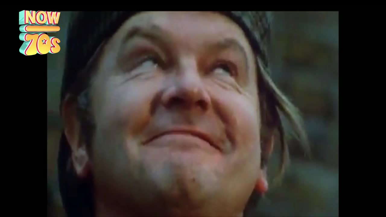 A man's face fills the frame, his eyes rolling upwards with a wide, almost mischievous grin. The grainy footage, reminiscent of 1970s British television, captures a moment of pure, unadulterated mirth.