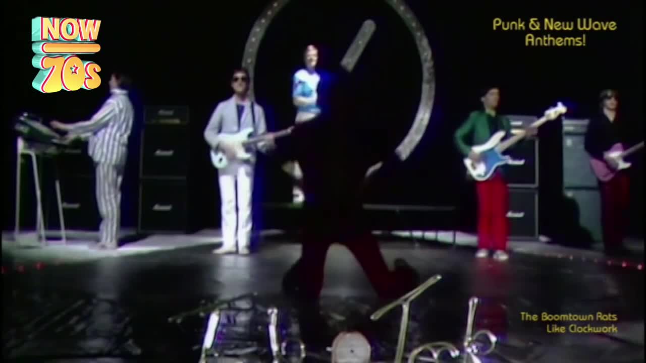 A band plays on a dark stage, bathed in spotlights. The keyboardist wears striped pajamas, while a guitarist in a pale suit stands beside him.