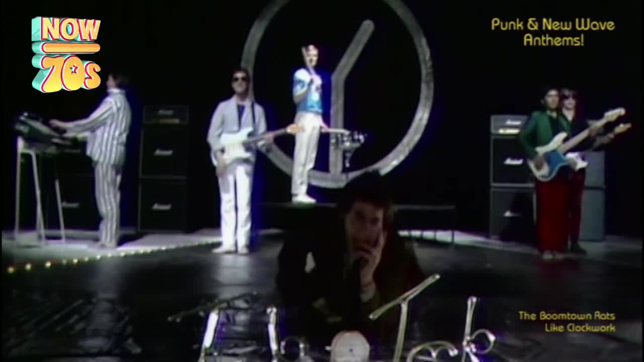 A keyboardist in striped pajamas plays a synthesizer while a guitarist in a light suit stands beside him. The lead singer, in a blue and white top, stands on a raised platform, holding a microphone.