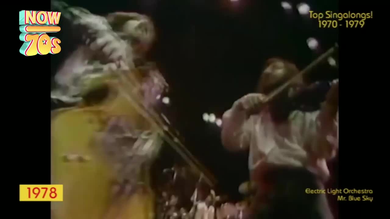 A man in a white shirt plays the violin, his bow moving across the strings. Another musician, blurred by motion, stands beside him, the stage lights glinting off their instruments.