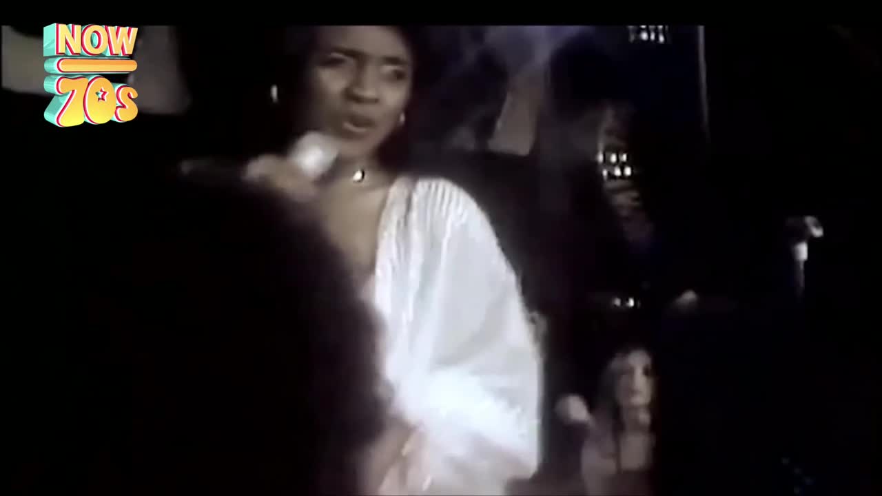 A woman in a white flowing dress sings into a microphone, her expression intense. The stage lights catch her face, illuminating her as she performs.