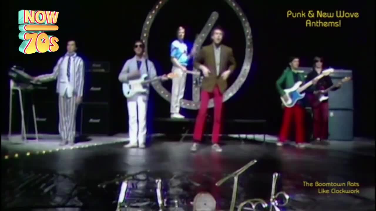 The Boomtown Rats are on stage, bathed in light. Bob Geldof, in a brown jacket and bright red trousers, bounces on the balls of his feet.