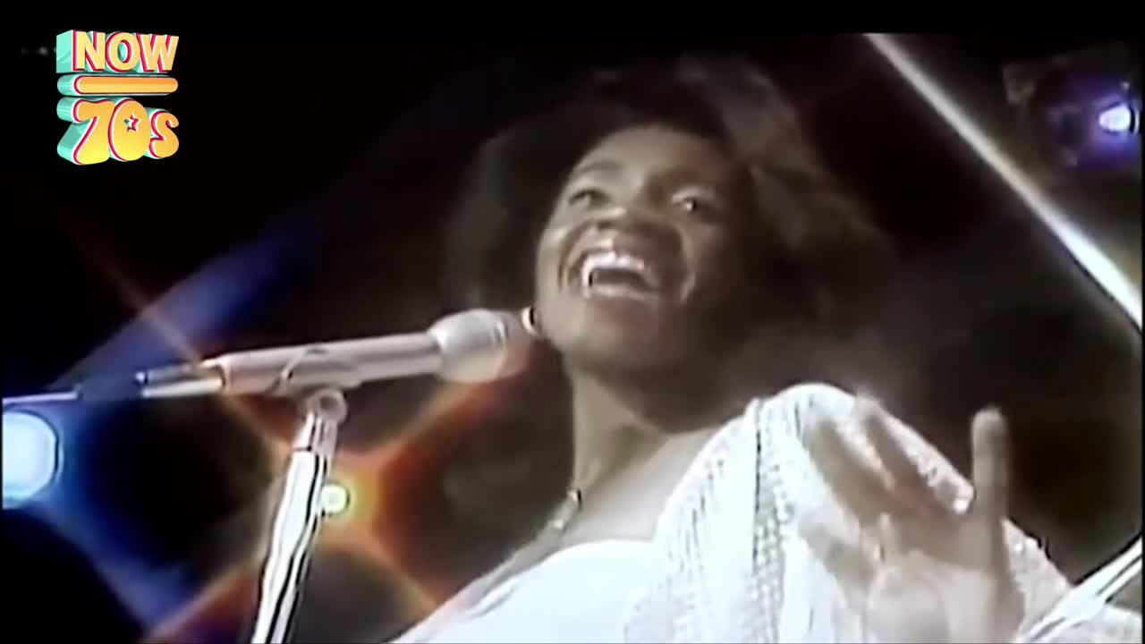 A woman belts into a microphone, her face alight with joy. Her white dress shimmers under the stage lights.
