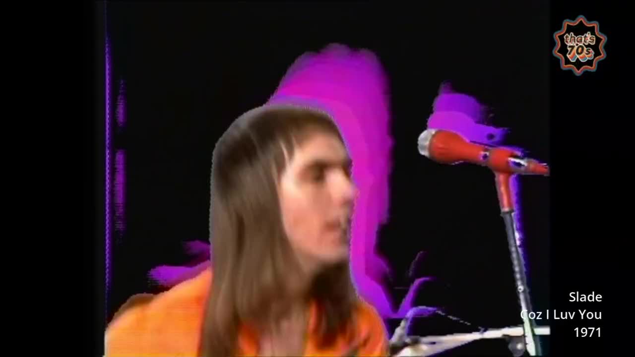 A young man with long brown hair, wearing an orange shirt, moves his head from side to side. A bright red microphone on a stand is positioned to his right.