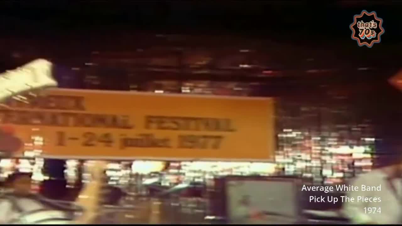 A bright orange banner for the "International Festival 1-24 July 1977" passes by. Lights from the stage and a crowd are visible behind it.