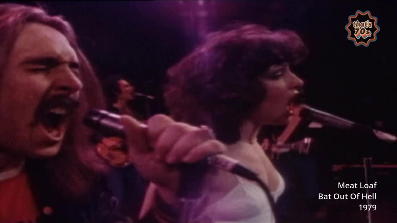 A man with a thick mustache belts into a microphone, his mouth wide open. Beside him, a woman with dark, curly hair also sings into her mic, her eyes closed.