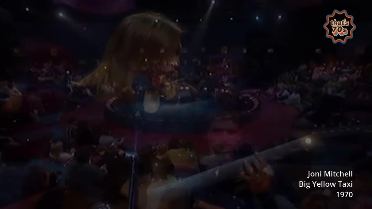 Joni Mitchell is on stage, her blonde hair catching the spotlights as she strums her guitar. The audience is a sea of faces in the dim theatre, all looking towards the performer.