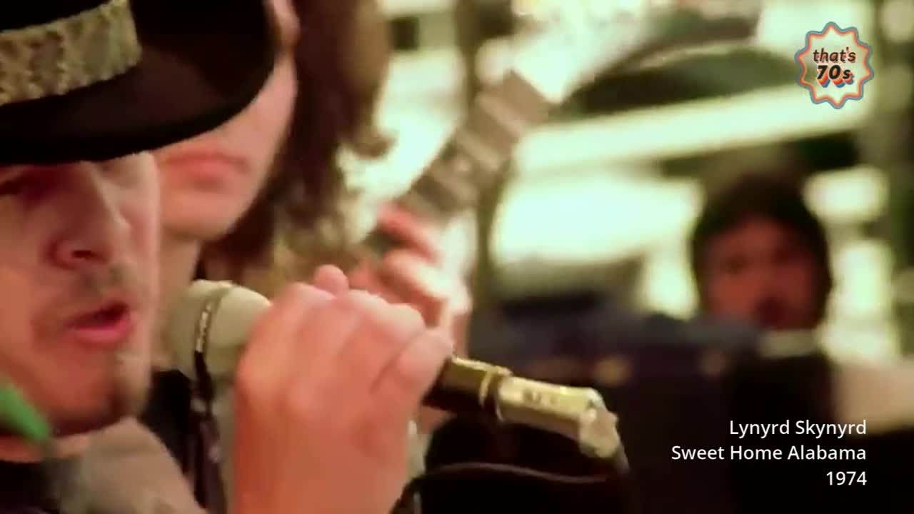 A man in a black hat sings into a microphone, his hand gripping the stand. Behind him, another musician strums a guitar.