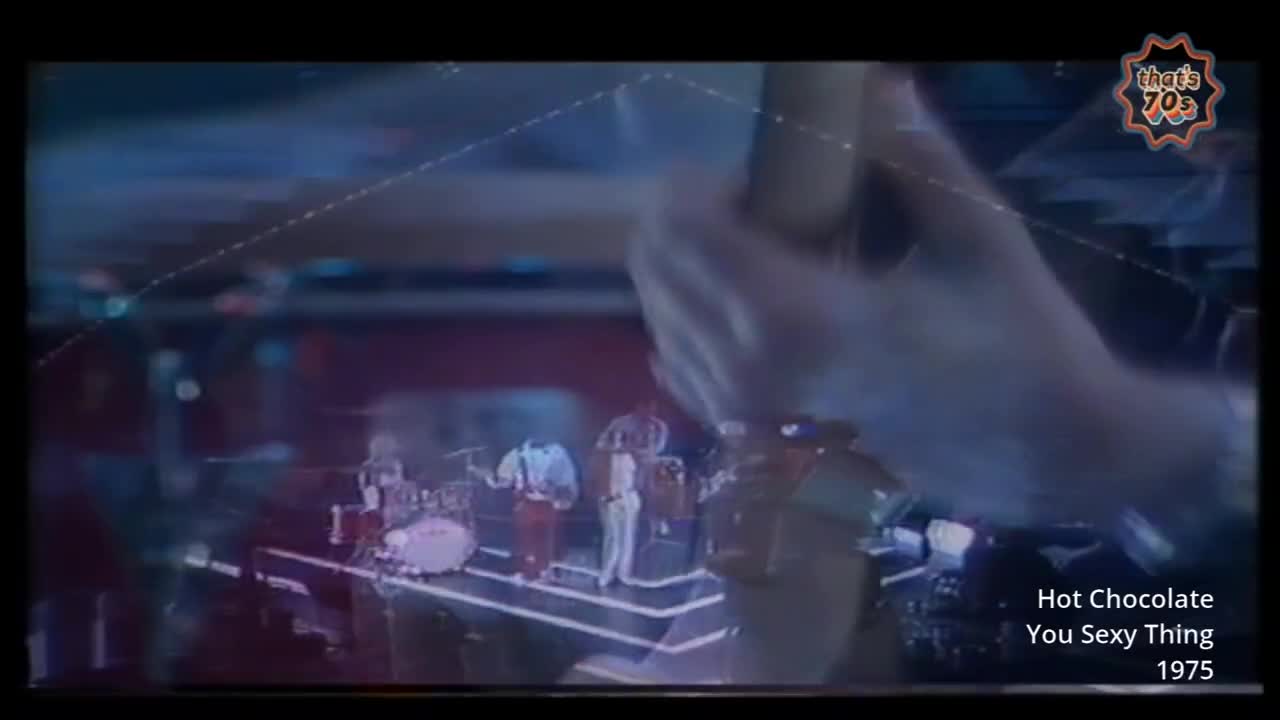 A band plays on a raised stage, bathed in blue and purple lights. The drummer hits his cymbal with a flourish while the singer, in a white suit, moves to the beat.