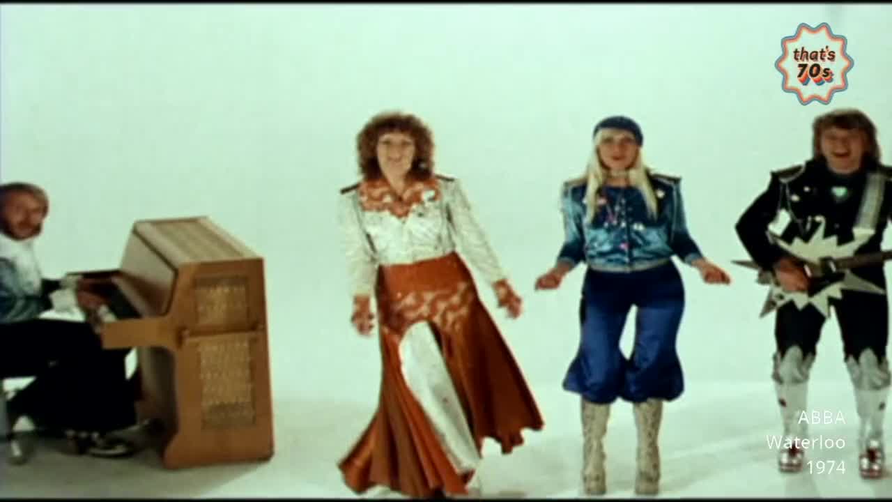 A man plays a wooden piano while two women in sparkly outfits dance. Another man strums a guitar with a starburst design.
