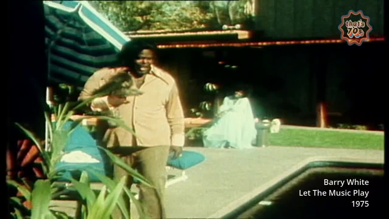 Barry White stands holding a bird, a light brown shirt and trousers on. A woman in a white dress is visible in the background.