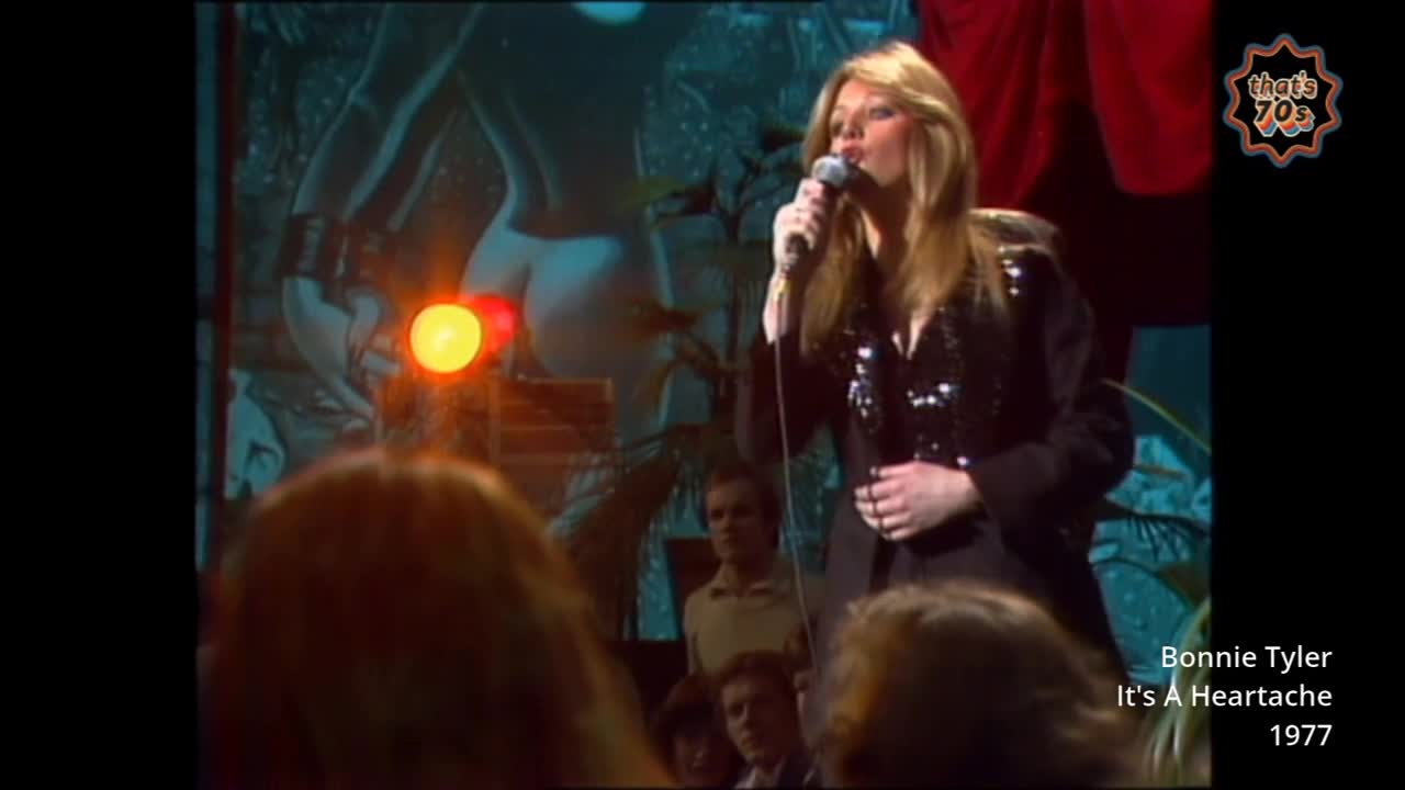 Bonnie Tyler stands centre stage, microphone in hand, her sequined jacket catching the light. A large, abstract backdrop featuring a stylized female form looms behind her.