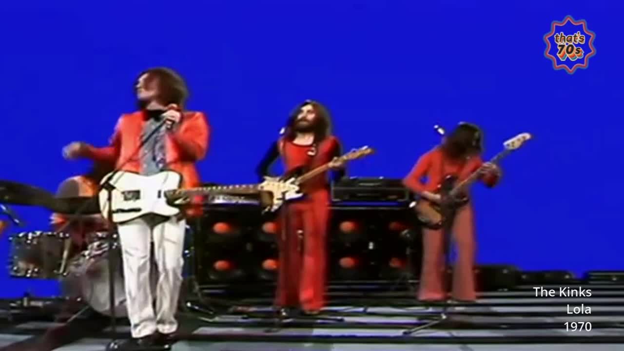 A band in bright red jumpsuits plays on a stage with a vibrant blue backdrop. The lead singer, in a red jacket and white trousers, holds a white guitar and sings into a microphone.