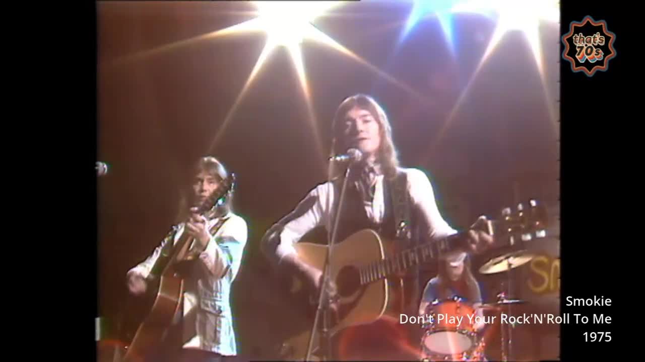 A man with long hair strums an acoustic guitar, singing into a microphone while bright stage lights beam down. Another guitarist stands to his left, also playing.