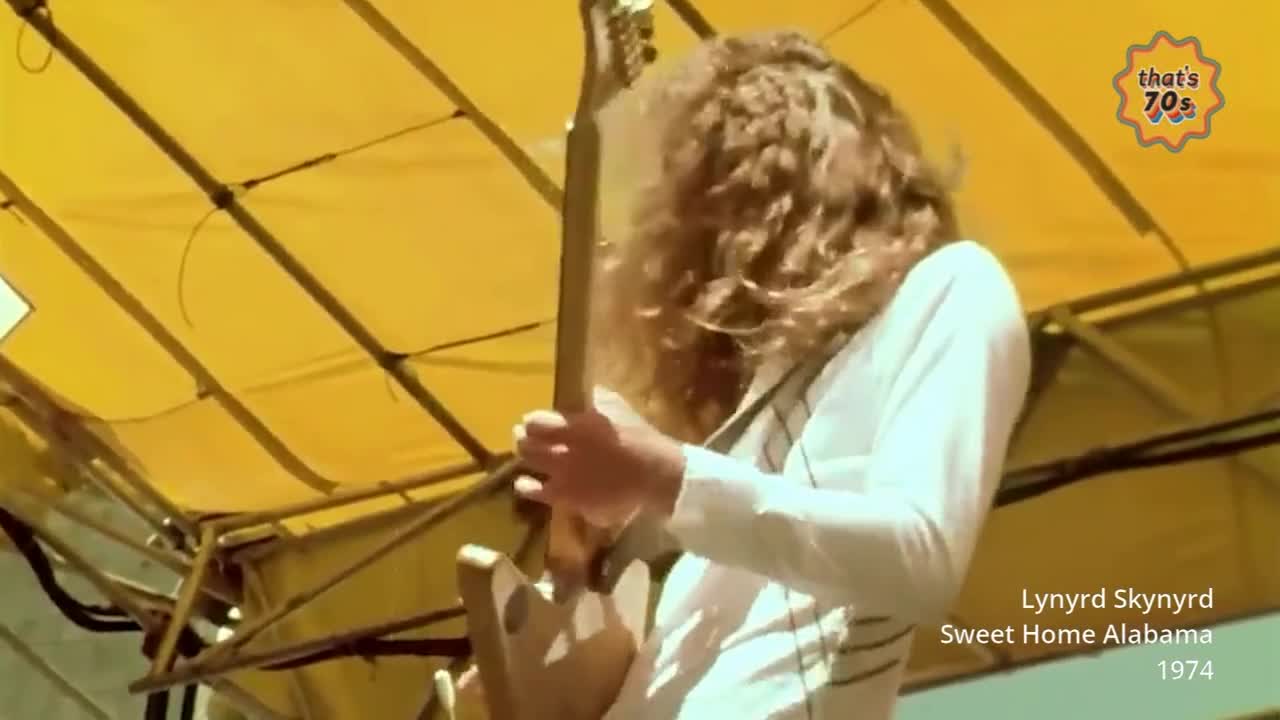 A guitarist with long, curly hair plays a sunburst electric guitar. He wears a white shirt with thin dark stripes under a yellow canopy.
