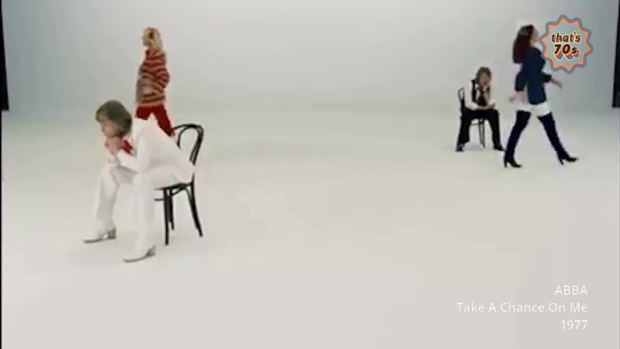 A woman in a white jumpsuit sits hunched on a chair, her hands clasped to her face. Across the stark white space, another woman in a striped top walks away, her red trousers a bold contrast.