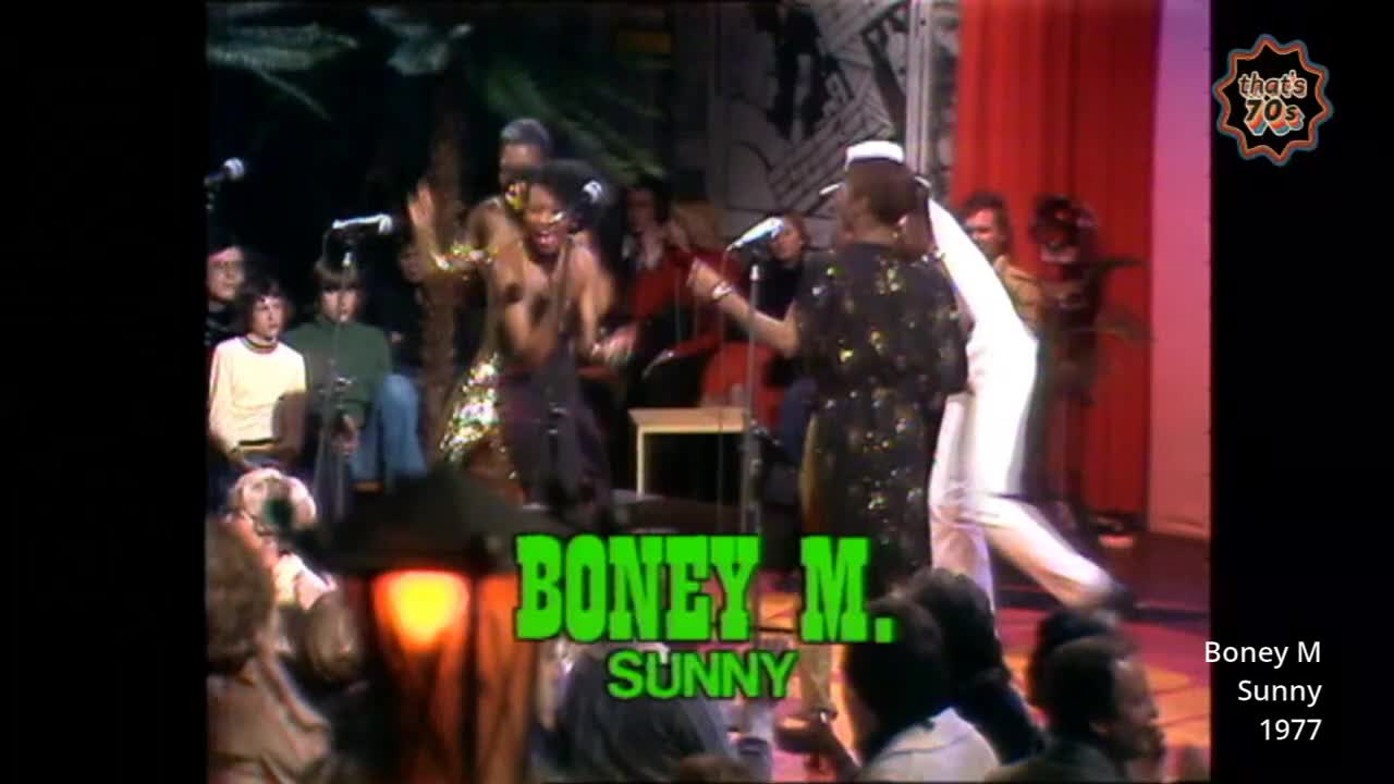 The lead singer, in a sparkly dress, is belting out a tune while her bandmates sway. A man in a white suit is dancing energetically behind her.