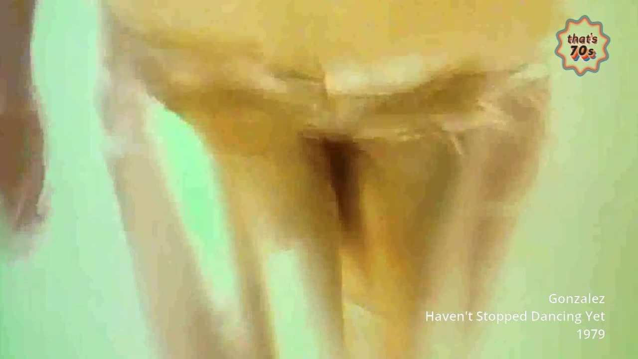 The legs of a dancer move in unison, their golden costume shimmering under the lights. The stage floor, a pale green, reflects the movement as the performer continues their routine.